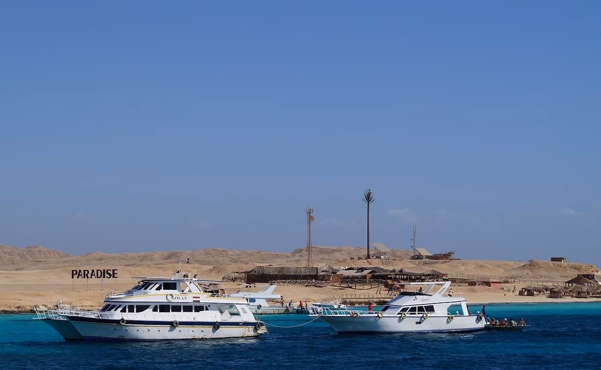 Twee witte excursieboten in diepblauw water voor een woestijneiland met strandhutten in de Rode Zee bij Hurghada