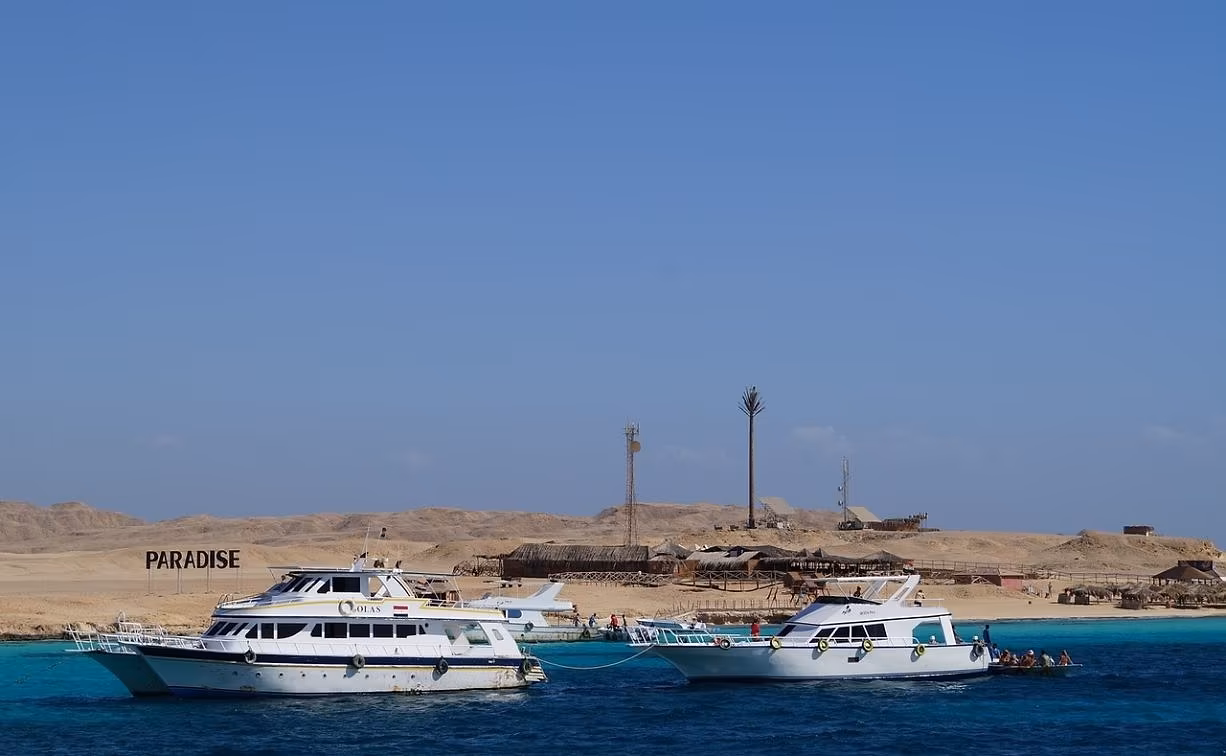 Twee witte excursieboten in diepblauw water voor een woestijneiland met strandhutten in de Rode Zee bij Hurghada