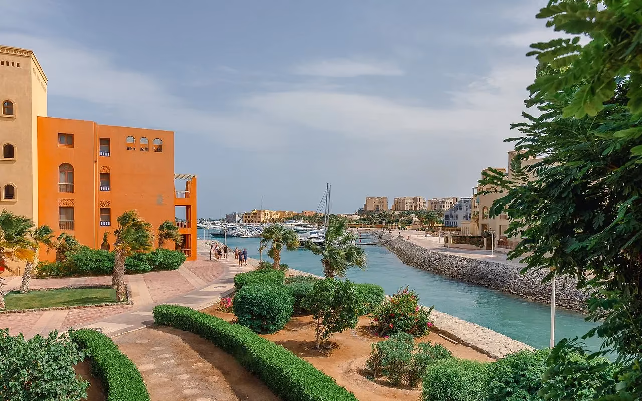 Een pad tussen heggen naar een oranje gebouw bij het water van een jachthaven in El Gouna