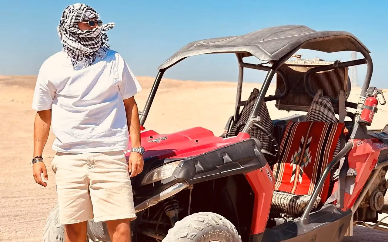 Een man met een geruite hoofddoek naast een rode buggy op zand in de woestijn onder een blauwe lucht