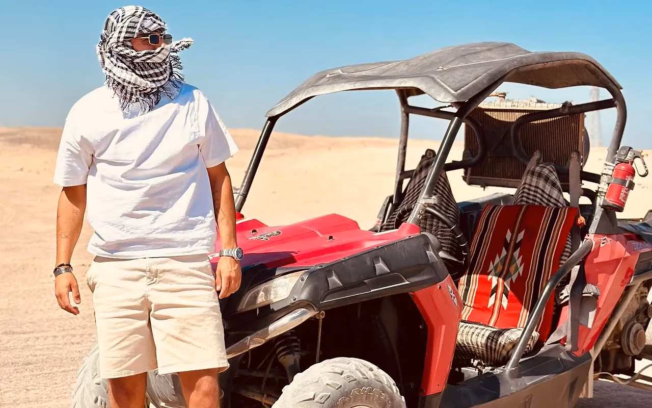 Een man met een geruite hoofddoek naast een rode buggy op zand in de woestijn onder een blauwe lucht