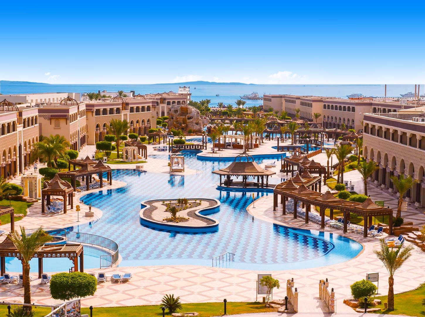 Overzicht van de lagunezwembaden met eilandjes, pergola's en palmbomen tussen de gebouwen in paleisstijl, met de Rode Zee aan de horizon, bij Sentido Mamlouk Palace Resort in Hurghada