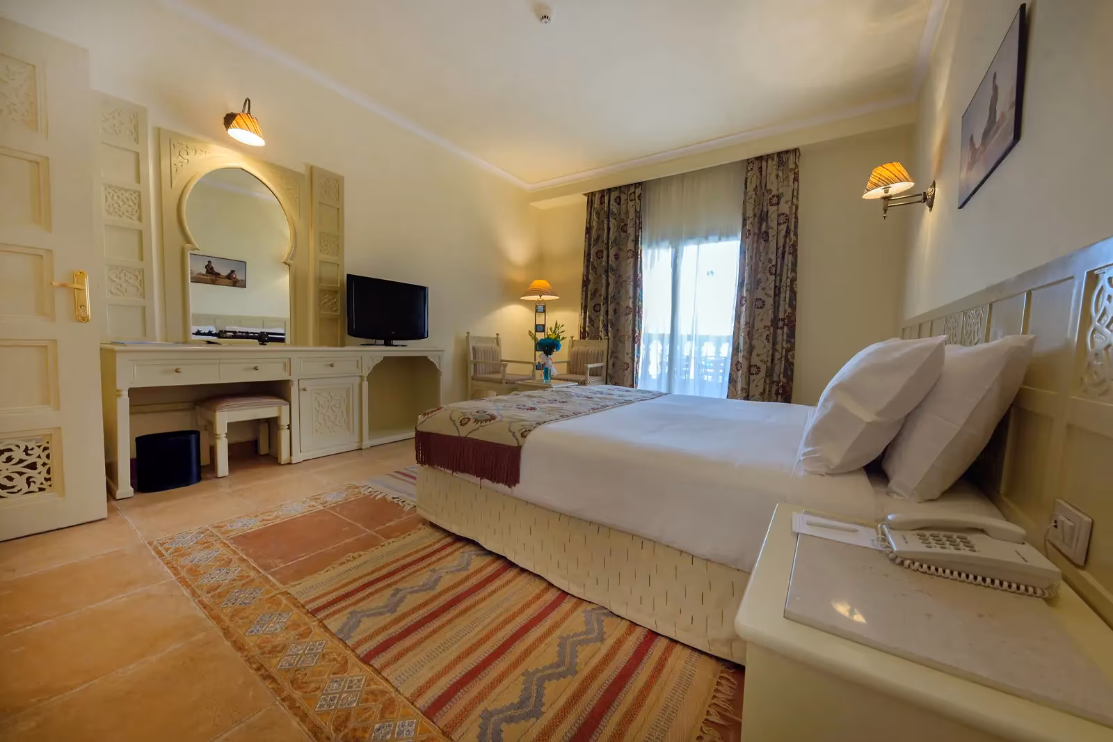 Slaapkamer met een tweepersoonsbed, een kaptafel met boogspiegel, een televisie en twee stoelen voor de balkondeur bij Sentido Mamlouk Palace Resort in Hurghada