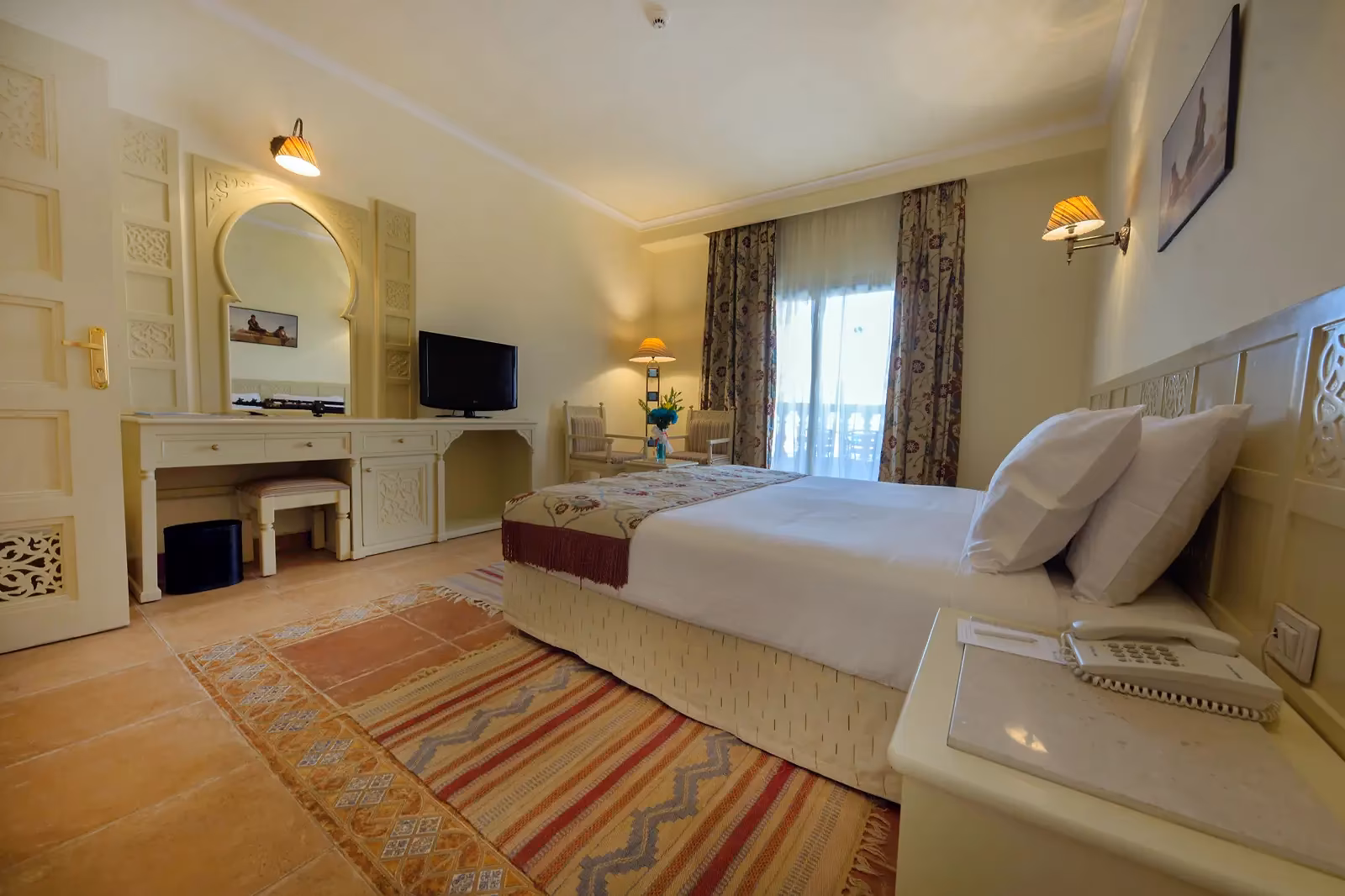Slaapkamer met een tweepersoonsbed, een kaptafel met boogspiegel, een televisie en twee stoelen voor de balkondeur bij Sentido Mamlouk Palace Resort in Hurghada