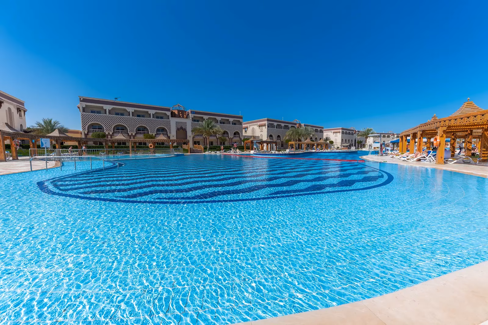Groot zwembad met een golvend tegelpatroon in donkerblauw, ligbedden en een houten paviljoen voor de gebouwen met bogen van Sentido Mamlouk Palace Resort in Hurghada