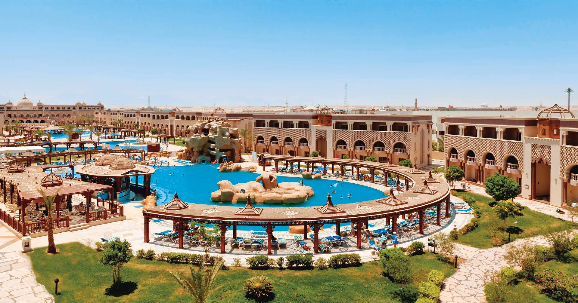 Rond zwembad met een rotseiland en glijbanen, omringd door pergola's, een gazon met palmbomen en gebouwen met bogen, met de zee in de verte, bij Sentido Mamlouk Palace Resort in Hurghada