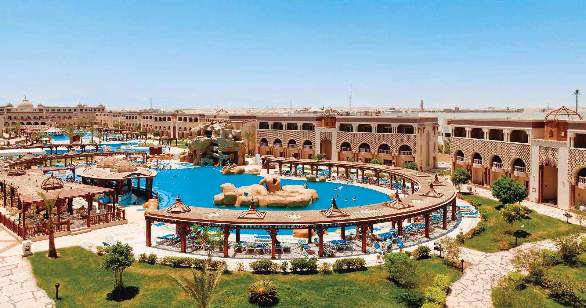 Rond zwembad met een rotseiland en glijbanen, omringd door pergola's, een gazon met palmbomen en gebouwen met bogen, met de zee in de verte, bij Sentido Mamlouk Palace Resort in Hurghada