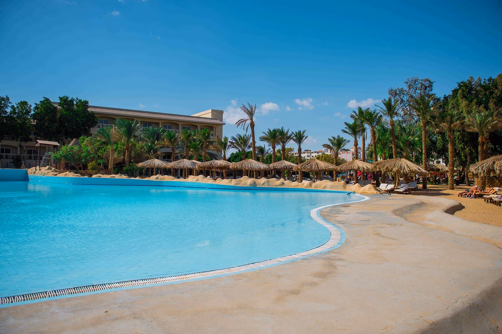 Lagunezwembad met een zandkleurige rand, rieten parasols en palmen voor een hotelgebouw bij Sindbad Club in Hurghada