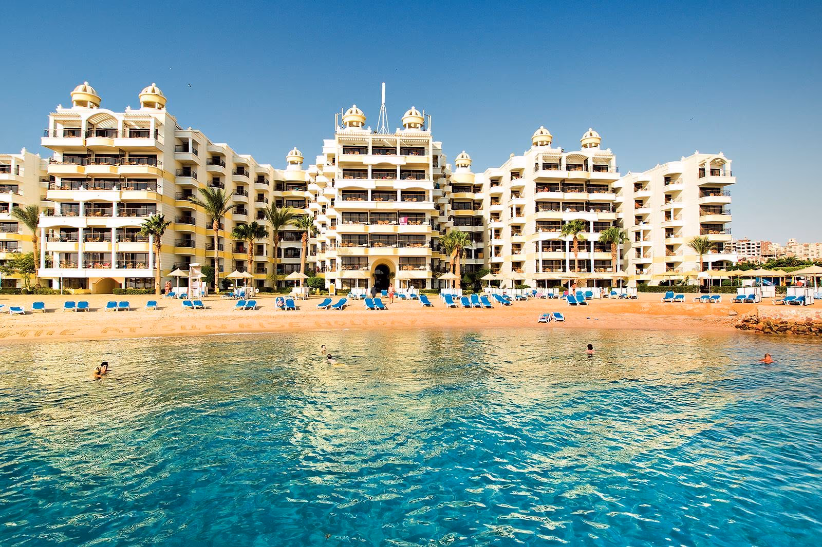 Witte hotelgebouwen met koepeltjes aan een baai met zandstrand, blauwe ligbedden en helder water bij Sunrise Holidays Resort in Hurghada